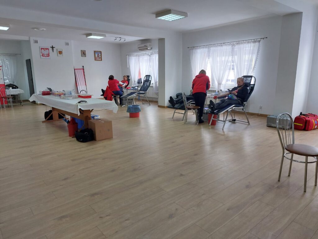 Pokój z jasną podłogą, białe ściany, dwie osoby leżące na fotelach medycznych, dwie osoby w czerwonych bluzach przy nich, stół z białym obrusem i sprzętem medycznym.