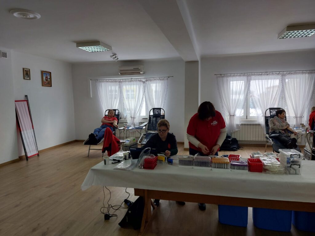 Pomieszczenie z jasnymi ścianami i drewnianą podłogą, trzy osoby na leżakach, dwie osoby przy stole z medycznymi probówkami i sprzętem, białe firanki na oknach.