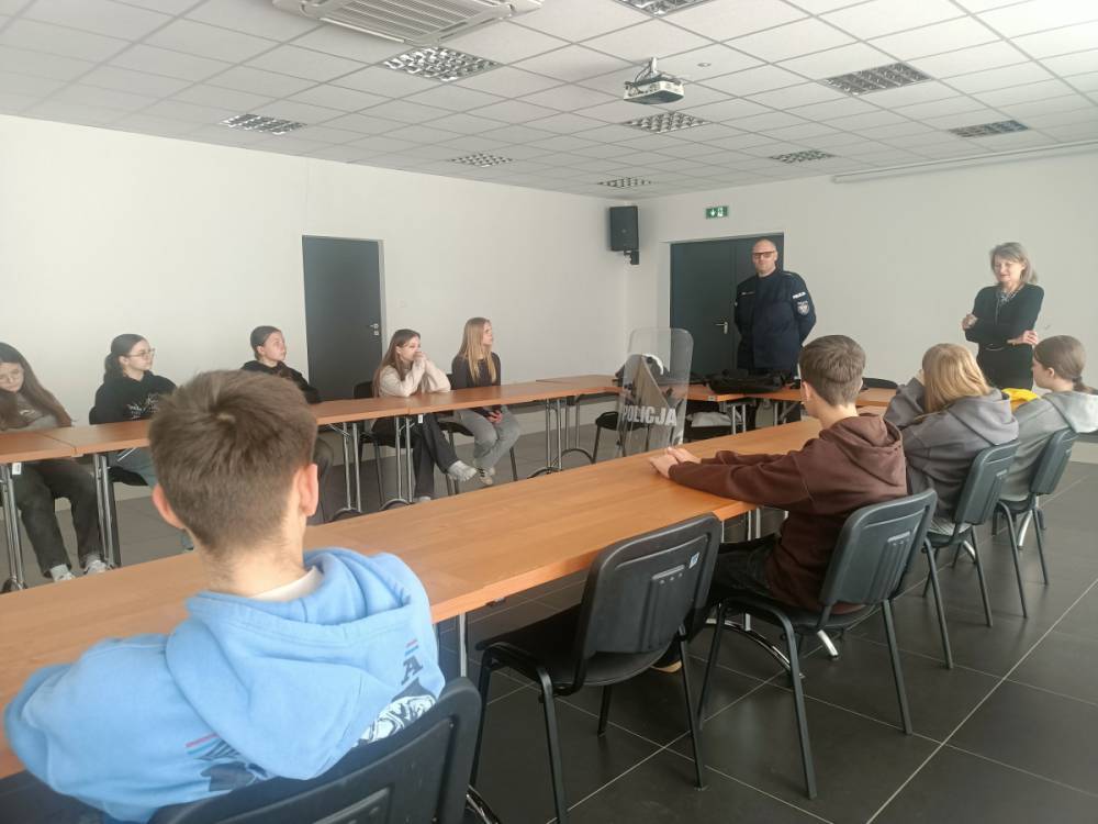 Grupa młodych osób siedzi przy stołach w sali konferencyjnej, dwie stojące osoby obok drzwi, na stole tarcza z napisem "Policja".