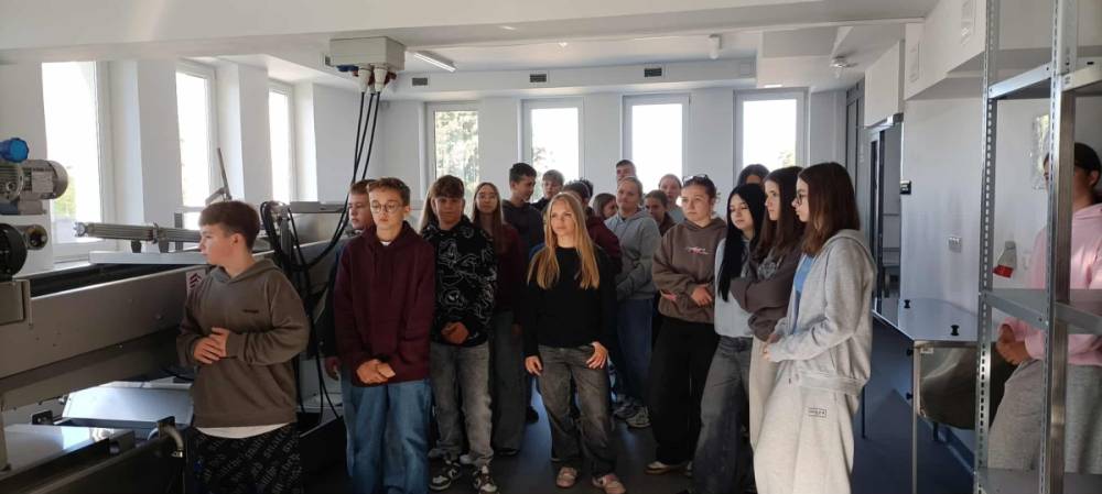 Grupa młodych osób stojących w jasnym pomieszczeniu z dużymi oknami i metalowymi urządzeniami po bokach.
