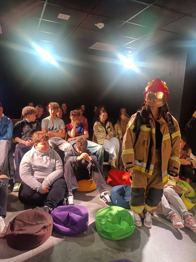 Grupa młodych osób siedzących na podłodze i pufach w ciemnym pomieszczeniu, jedna osoba stoi w stroju strażaka z czerwonym hełmem i żółto-brązowym ubraniem ochronnym.