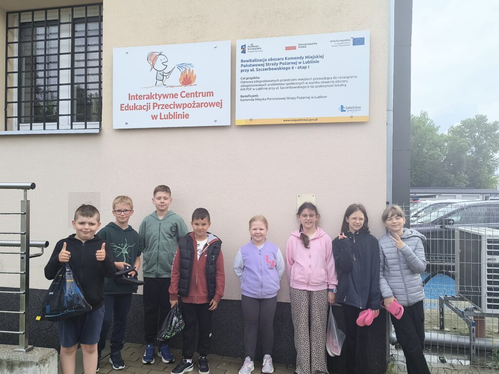 Grupa ośmiu dzieci stoi na tle jasnej ściany budynku z dwoma tablicami informacyjnymi, po prawej stronie ogrodzenie i zaparkowane samochody.