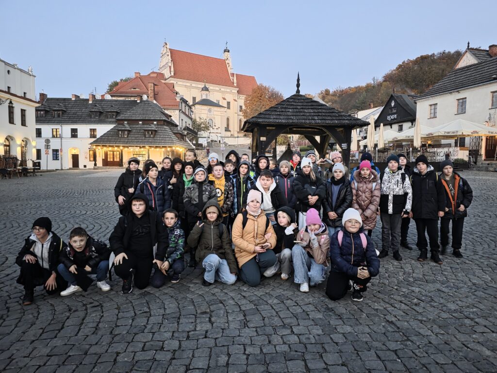 Grupa dzieci w kurtkach i czapkach na kamiennej nawierzchni na tle drewnianej altany i jasnych budynków z czerwonymi dachami.