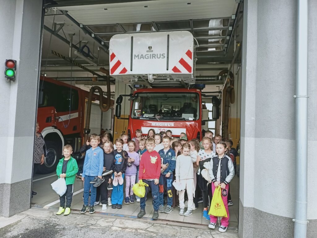 Grupa dzieci w kolorowych ubraniach stoi w garażu przed czerwonym samochodem strażackim marki Magirus, z szarymi ścianami i zielonym światłem sygnalizacji.