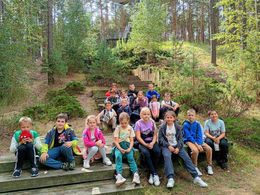 Grupa dzieci siedzi na drewnianych schodach w lesie, otoczona zielonymi drzewami i krzewami, ubrana w kolorowe ubrania i sportowe buty.
