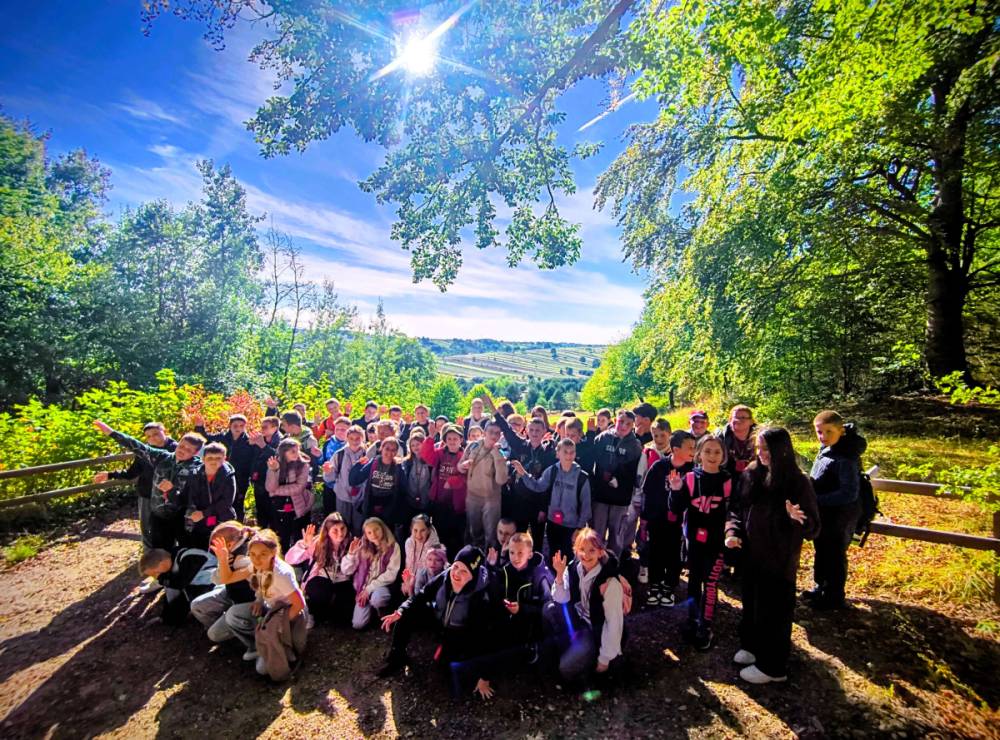 Grupa dzieci i młodzieży na leśnej ścieżce, wokół zielone drzewa, słoneczne niebo z jasnym słońcem, w tle pola i wzgórza.