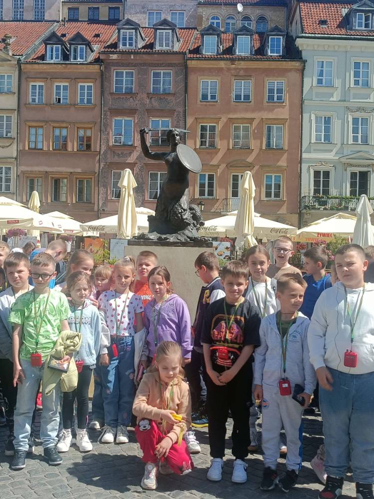 Grupa dzieci na tle pomnika syrenki i kolorowych kamienic z czerwonymi dachami oraz parasolami na rynku.