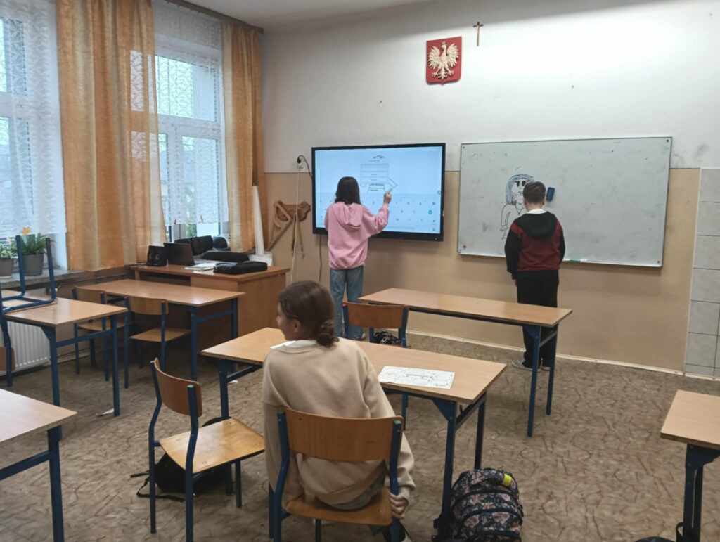 Klasa szkolna z jasnobrązowymi meblami, trzema uczniami, pracą na tablicy interaktywnej i białej, zasłony i rośliny przy oknie.