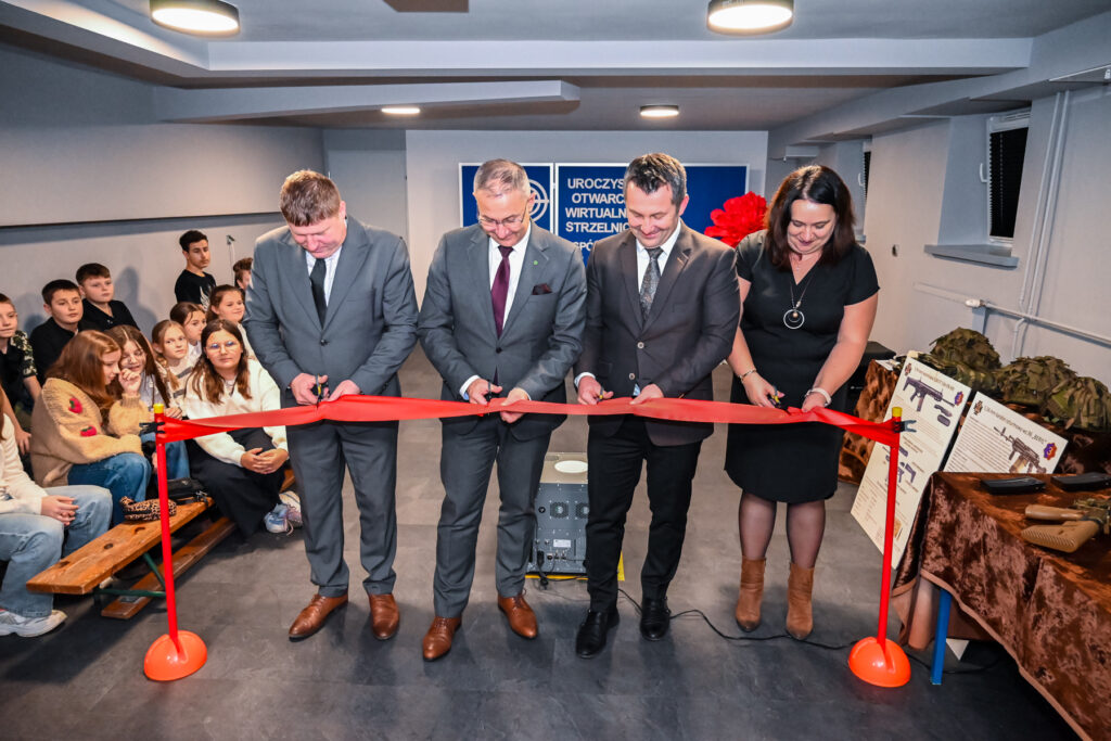 Cztery osoby w garniturach i sukience przecinające czerwoną wstęgę w pomieszczeniu, z lewej strony siedzi grupa dzieci, z prawej stoły z hełmami i planszami informacyjnymi.