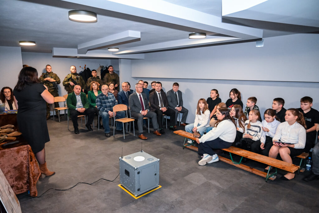 Grupa dorosłych i dzieci siedzących na krzesłach i ławkach w jasnym pomieszczeniu, kobieta w czarnej sukience stoi na tle stołu, na podłodze metalowa skrzynka z antenami.