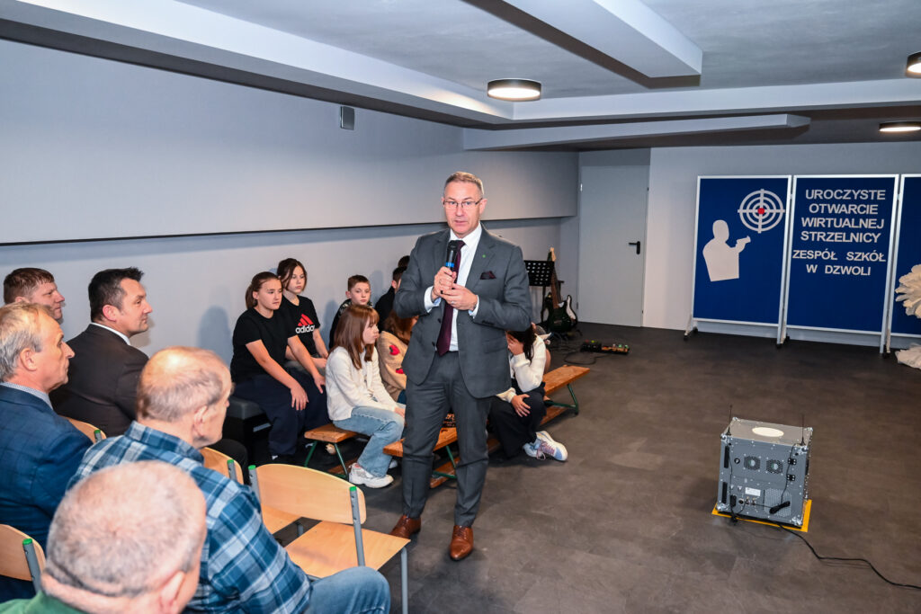 Mężczyzna w garniturze z mikrofonem stoi na sali, wokół niego siedzą dzieci i dorośli, na ścianie plansze z napisem „Uroczyste otwarcie wirtualnej strzelnicy”.