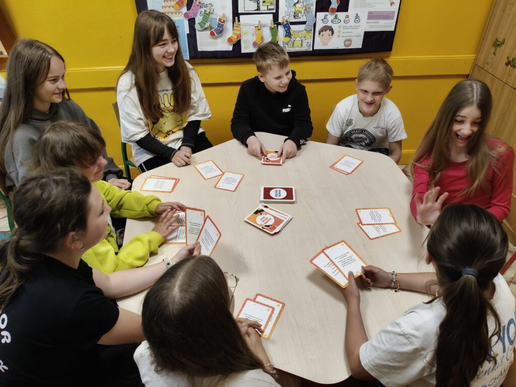 Grupa dzieci siedzi wokół jasnego, ośmiokątnego stołu, grając w grę karcianą, w tle żółta ściana z tablicą i kolorowymi dekoracjami.