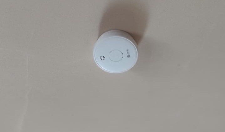 Biały okrągły czujnik dymu zamontowany na jasnoszarym suficie.