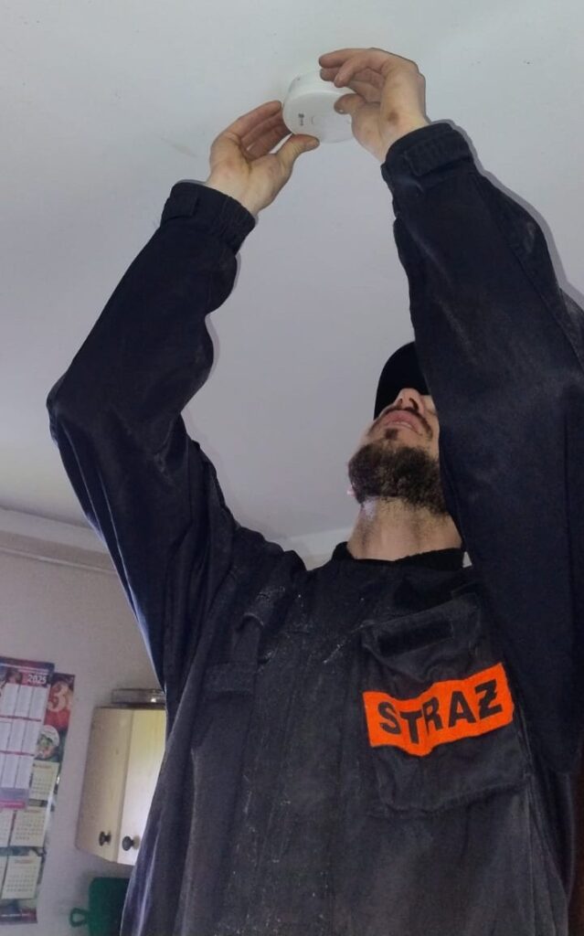 Mężczyzna w czarnej kurtce z napisem "STRAŻ" montuje biały czujnik na białym suficie w pomieszczeniu z kalendarzem i meblami.