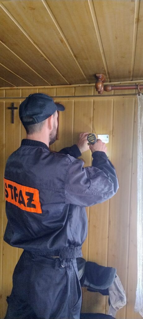 Mężczyzna w czapce i granatowym ubraniu z napisem "STRAŻ" na plecach wierci ścianę z jasnego drewna wewnątrz pomieszczenia.