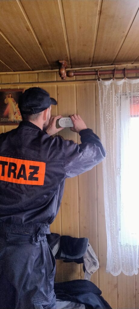 Mężczyzna w czarnej kurtce z napisem STRAŻ i czapce instaluje biały czujnik dymu na drewnianej ścianie w pokoju z białą koronkową zasłoną przy oknie.