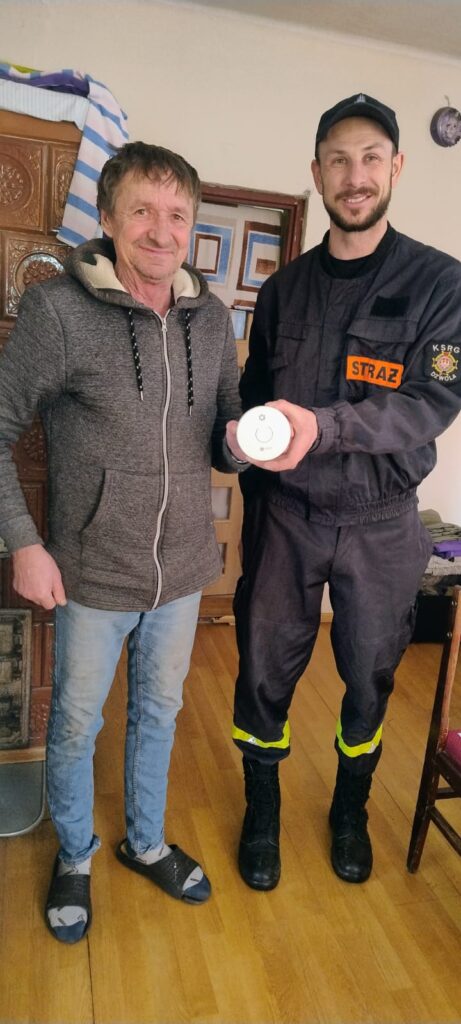 Dwóch mężczyzn stoi na drewnianej podłodze, jeden w szarej bluzie i jasnych spodniach, drugi w czarnym mundurze strażackim z pomarańczową naszywką "STRAŻ" trzyma biały okrągły przedmiot.