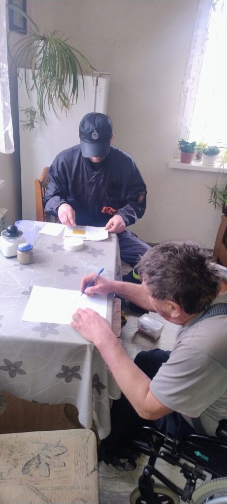 Dwóch mężczyzn siedzi przy stole z jasnym obrusem w kwiaty, jeden pisze długopisem na kartce, drugi trzyma dokument, w tle okno z kwiatami i roślina.