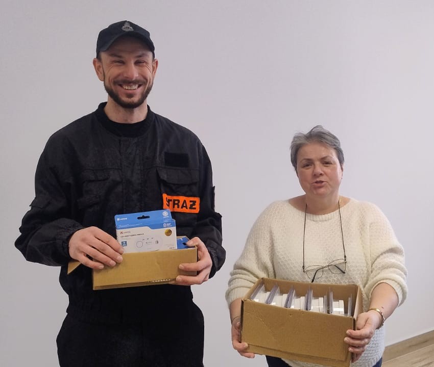 Mężczyzna w czarnym mundurze z napisem STRAŻ i kobieta w białym swetrze trzymają kartonowe pudełka z opakowaniami urządzeń elektronicznych na jasnym tle.