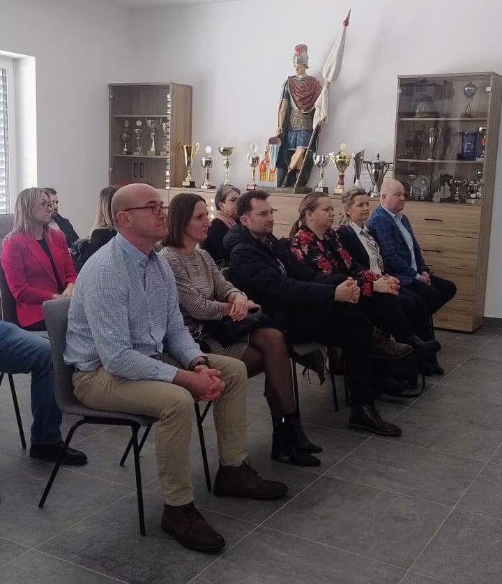 Grupa siedzących osób w rzędzie na szarych krzesłach, w tle szafki z pucharami i duża figurka rzymskiego żołnierza z proporcem.