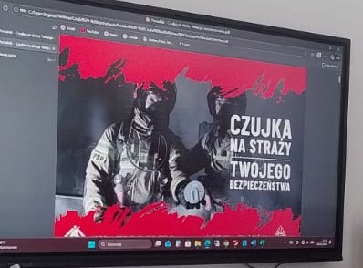 Plakat z dwoma strażakami w hełmach i maskach, czerwone plamy na górze i dole, biały tekst "Czujka na straży twojego bezpieczeństwa" na szarym tle.