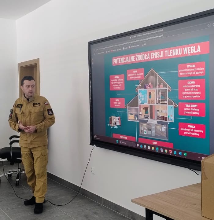 Mężczyzna w mundurze stoi obok dużego ekranu z infografiką domu i opisami potencjalnych źródeł emisji tlenku węgla, wnętrze jasnego pomieszczenia.
