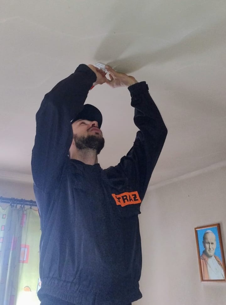 Mężczyzna w czarnej bluzie z napisem FRAZ montujący czujnik dymu na białym suficie w jasnym pomieszczeniu z zasłonami i obrazem na ścianie.